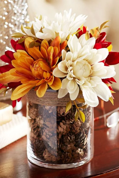 Herbstdeko mit Tannenzapfen großes Glas als Mittelstück auf dem Tisch gefüllt mit Kiefernzapfen mit Herbstblumen Chrysanthemen verziert
