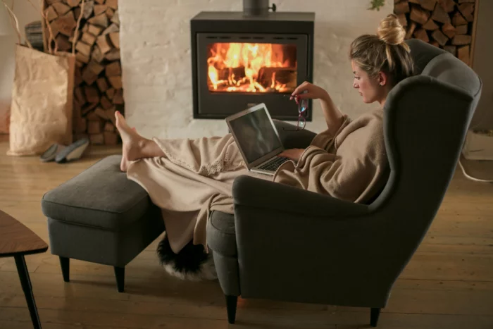 gesund und glücklich im Winter junge Frau im Sessel vor dem brennenden Kamin ein Buch in der Hand Brennholz warme gemütliche Raumatmosphäre