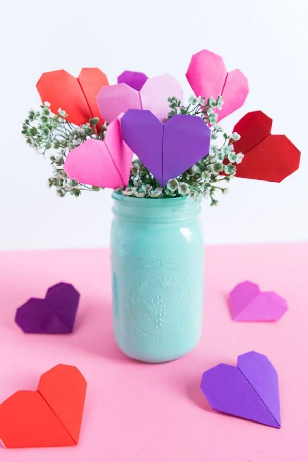 DIY Geschenke zum Valentinstag kleine Origami Herzen aus buntem Papier in einem hellblauen Einweckglas mit Blumen arrangiert