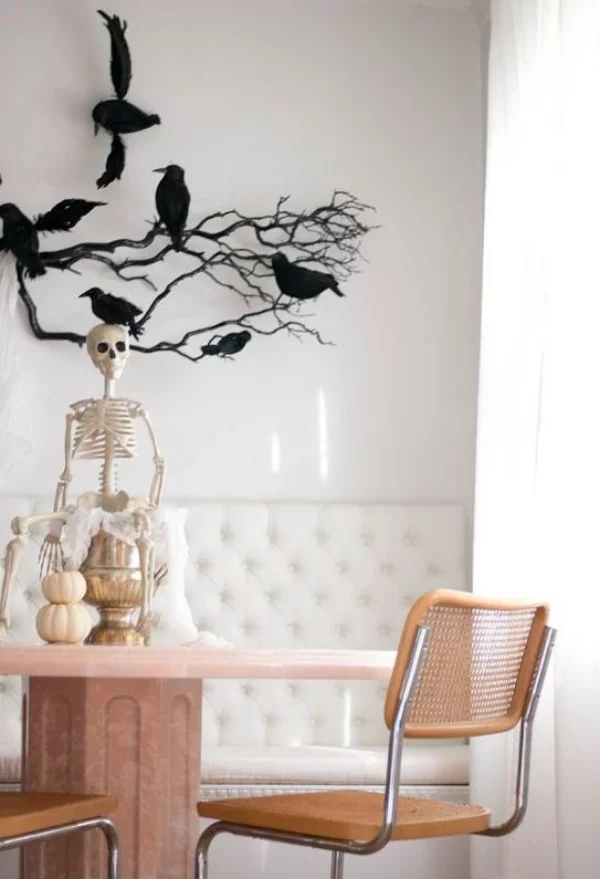 minimalistische Halloween Deko Essecke in hellen Farbtönen schwarze Zweige Raben an der Wand Skelett auf dem Tisch zwei kleine Kürbisse