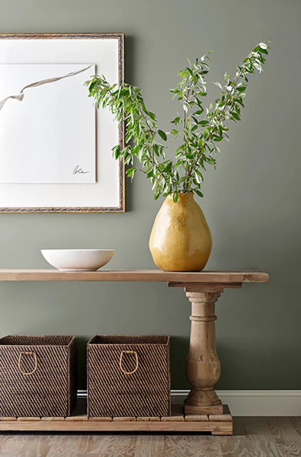 Evergreen Fog Farbe des Jahres 2022 Ruhe und Gelassenheit ausstrahlen Holztisch große Vase mit grünen Zweigen grau-grüne Wandfarbe Flechtkörbe